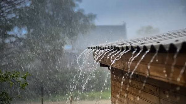 Das Bild zeigt starken Regen, der von einem Wellblechdach auf eine Holzhütte herabströmt.