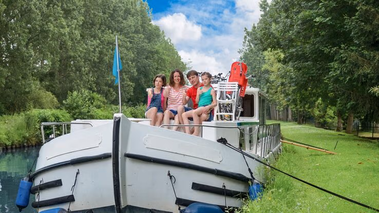 Das Foto zeigt vier Personen auf einem Hausboot. Sie sitzen an der Front des Bootes und umarmen sich. Das Boot ist noch mit Seilen an Pfosten festgemacht.
