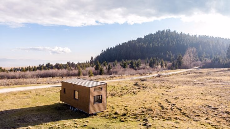 Das Foto zeigt ein Caravan auf einem Feld. Es ist ein kleiner Wohnwagen aus Holz der auf Rädern steht. Im Hintergrund ist ein Hügel mit einem Wald zu sehen.