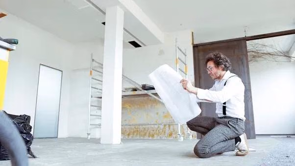 Ein Mann hält einen Bauplan in der Hand und kniet dabei auf dem Boden.