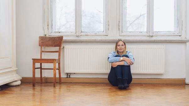 Person sitzt in einem hellen, weitgehend leeren Wohnraum mit Holzfußboden vor einer Fensterfront und einem Heizkörper.