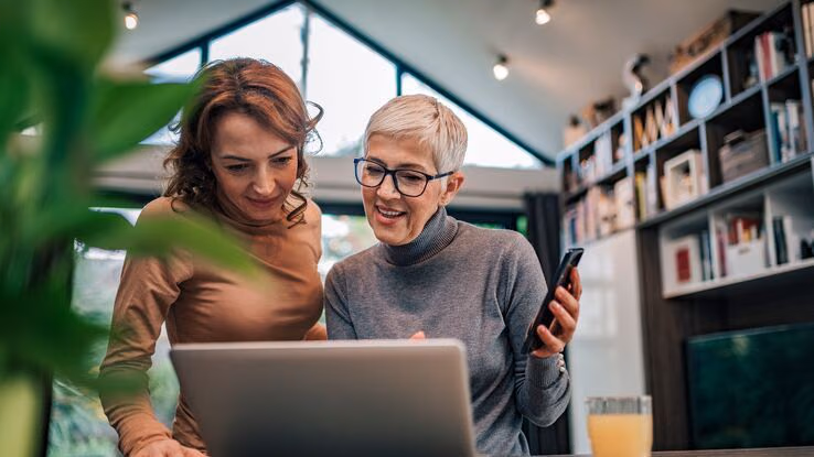 Zwei Frauen informieren sich an ihrem Laptop über ein Thema. Sie wirken zufrieden und konzentriert.