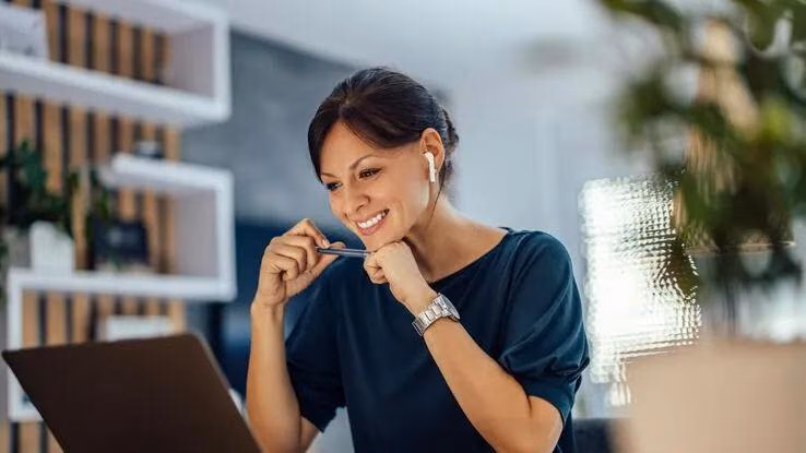 Eine Frau lächelt breit in ihren Laptop, während sie einen Stift in den Händen hält.