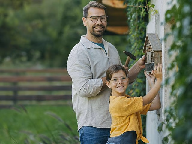 Ein Vater bringt mit seiner Tochter ein Vogelhaus an der Hauswand seines Eigenheims an.