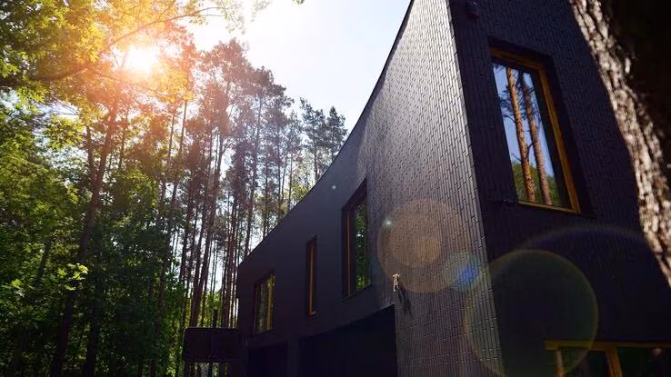 Das Foto zeigt eine schwarze Villa, die neben einem Wald steht. Sie ist leicht gebogen und hat große Fenster. Durch die Bäume ist das Haus von der Sonne geschützt.