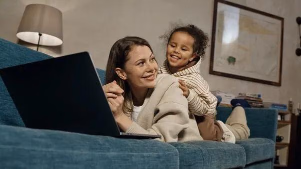 Eine Mutter liegt auf dem Bauch auf dem Sofa und informiert sich über Kaufnebenkosten. Ihre Tochter sitzt auf ihr drauf.