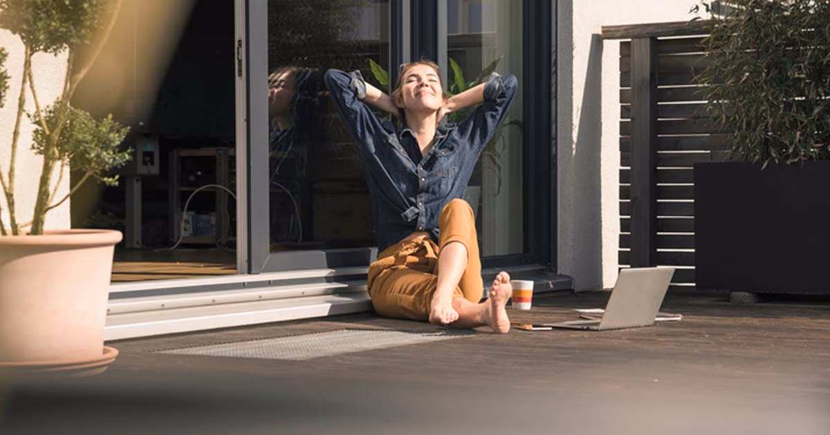 Eine Frau sitzt auf ihrer Terrasse mit einem Laptop und genießt die Sonne.