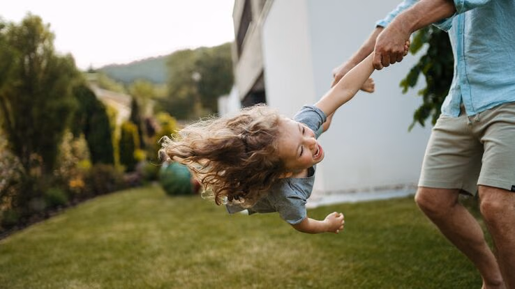 Ein Vater nimmt Fuß und Hand seiner Tochter und schwingt sie durch den Garten.