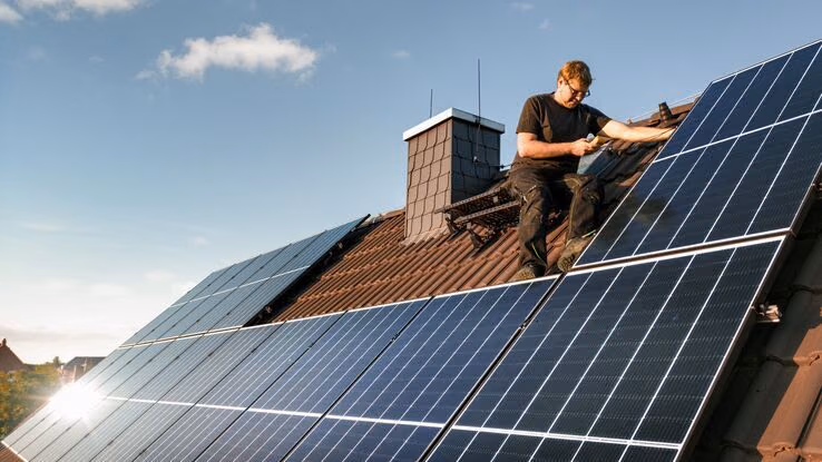 Ein Handwerker befindet sich auf einem Dach und montiert Photovoltaikmodule. Er trägt Arbeitskleidung und ist mit der Installation der Solaranlage beschäftigt.