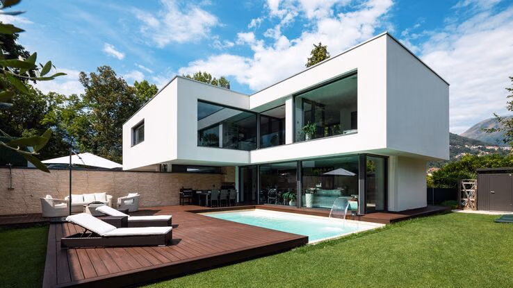 Abgebildet ist ein sehr modernes, weißes Haus mit großen Fenstern und einem eingebauten Pool auf der Gartenterasse.