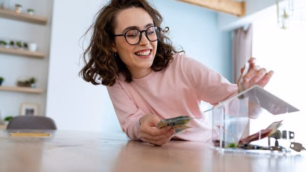 Eine Frau wirft Geld in eine Spardose, die aussieht wie ein Haus.