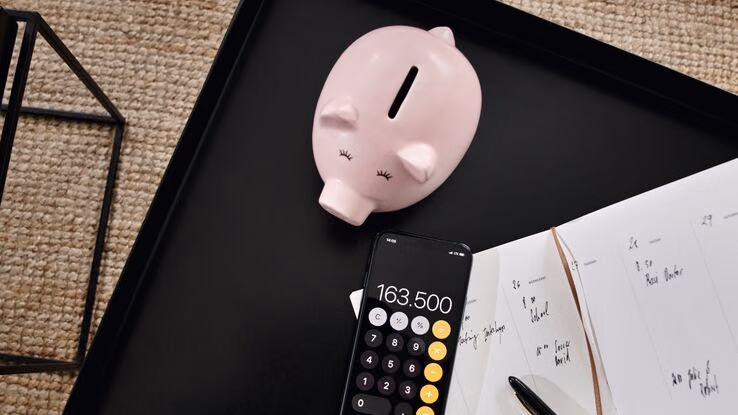 Auf einem schwarzen Tisch liegen ein rosa Sparschwein, ein Smartphone mit geöffneter Rechner-App sowie ein Notizbuch mit handschriftlichen Einträgen. Daneben liegt ein Stift.