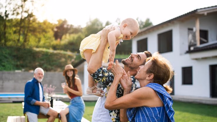 Das Foto zeigt eine Familie in einem schönen Garten. Im Vordergrund hebt der Vater seine Tochter in die Luft und die Großmutter steht daneben.