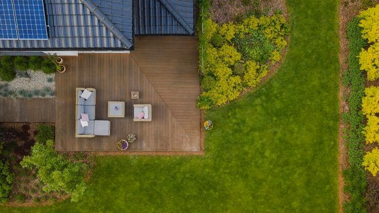 Es ist ein Haus mit Photovoltaik und einer großen Terasse und einem Garten mit einer großen Fläche an Wiese aus der Vogelperspektive abgebildet.