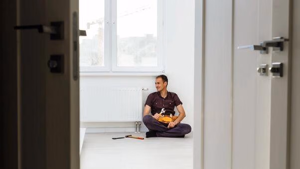 Ein Handwerker sitzt an der Heizung. Das Bild von der Tür aus gemacht, weshalb diese an den Seiten noch zu erkennen ist.