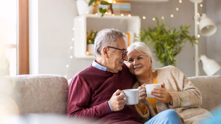 Ein älteres Pärchen sitzt auf dem Sofa und trinkt Kaffee. Die Frau kuschelt sich an den Mann.