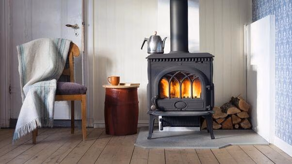 Ein altmodischer Kamin in dem ein Feuer brennt. Dahinter liegt ein Haufen Holzscheite. Neben dem Kamin stehen ein Stuhl mit einer Decke und ein Tischchen mit einer Tasse darauf.