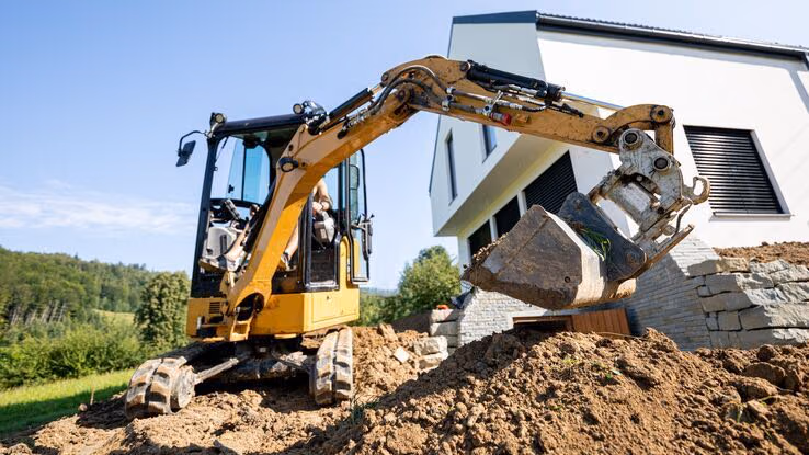 Im Vordergrund sieht man einen Bagger auf dem Grundstück und im Hintergrund ein modernes Neubauhaus.